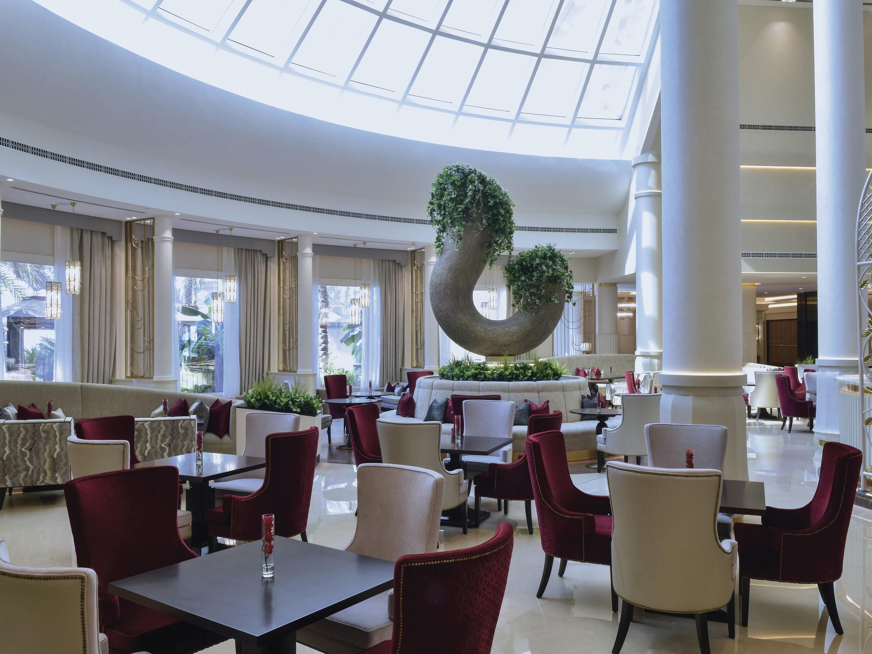 A spacious hotel lobby with high ceilings, large windows, and a central decorative tree.