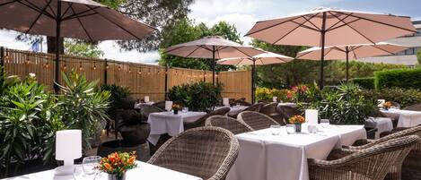 An outdoor dining area with wicker chairs and tables, covered by large umbrellas, and surrounded by greenery.