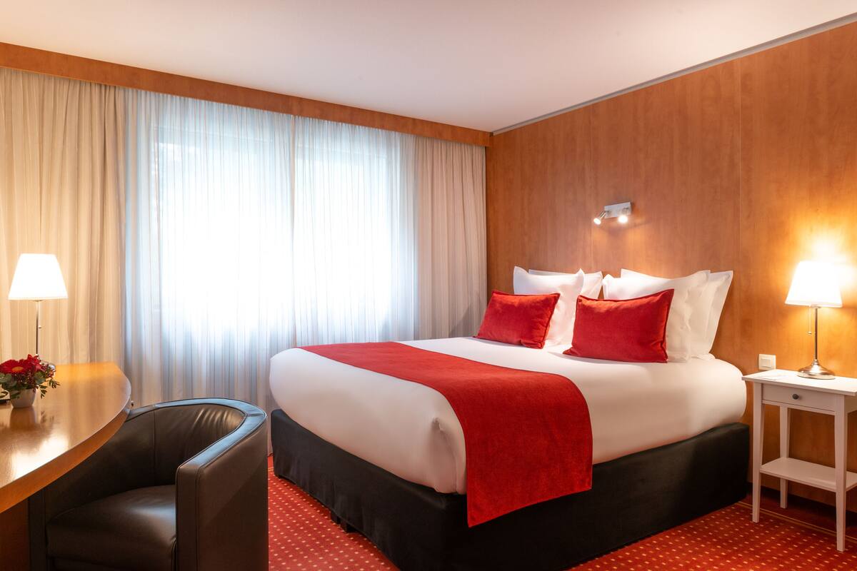 Habitación de hotel con una cama grande, un escritorio con silla, una mesita pequeña y una lámpara.