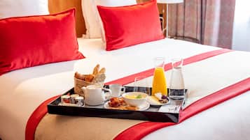 Una cama bien hecha con almohadillas rojas, una bandeja de desayuno con cruasanes, café, huevos y jugo, y una colcha a rayas rojas y blancas.
