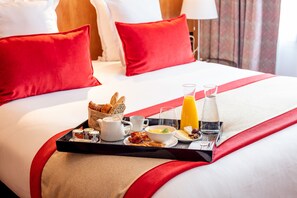 Una cama bien hecha con almohadillas rojas, una bandeja de desayuno con cruasanes, café, huevos y jugo, y una colcha a rayas rojas y blancas.