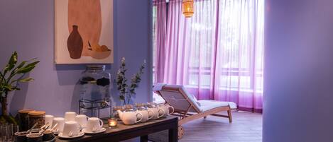 Una habitación con una chaise longue, una mesa con tazas y un florero, y un cuadro de un florero y una planta.