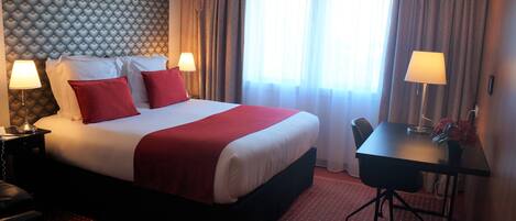Une chambre d’hôtel avec un grand lit, deux tables de chevet avec des lampes, un bureau avec une chaise et une fenêtre avec des rideaux.