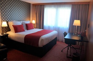 Quarto de hotel com uma cama grande, duas mesas de cabeceira com luminárias, uma escrivaninha com cadeira e uma janela com cortinas.
