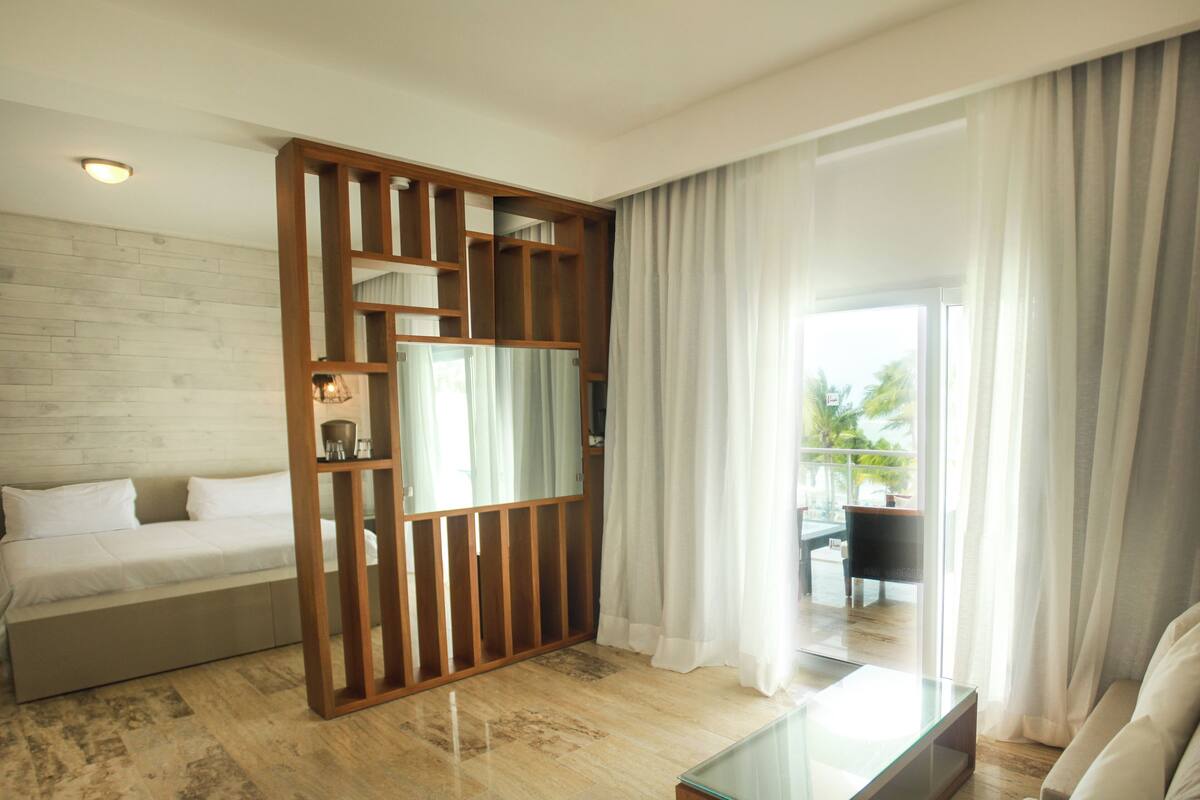 Ein modernes Hotelzimmer mit einer Holzwand, einer Couch, einem Glastisch und Blick auf einen Balkon mit Gartenmöbeln.
