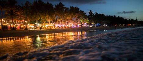 A beach at night with palm trees and a lit-up area in the background.