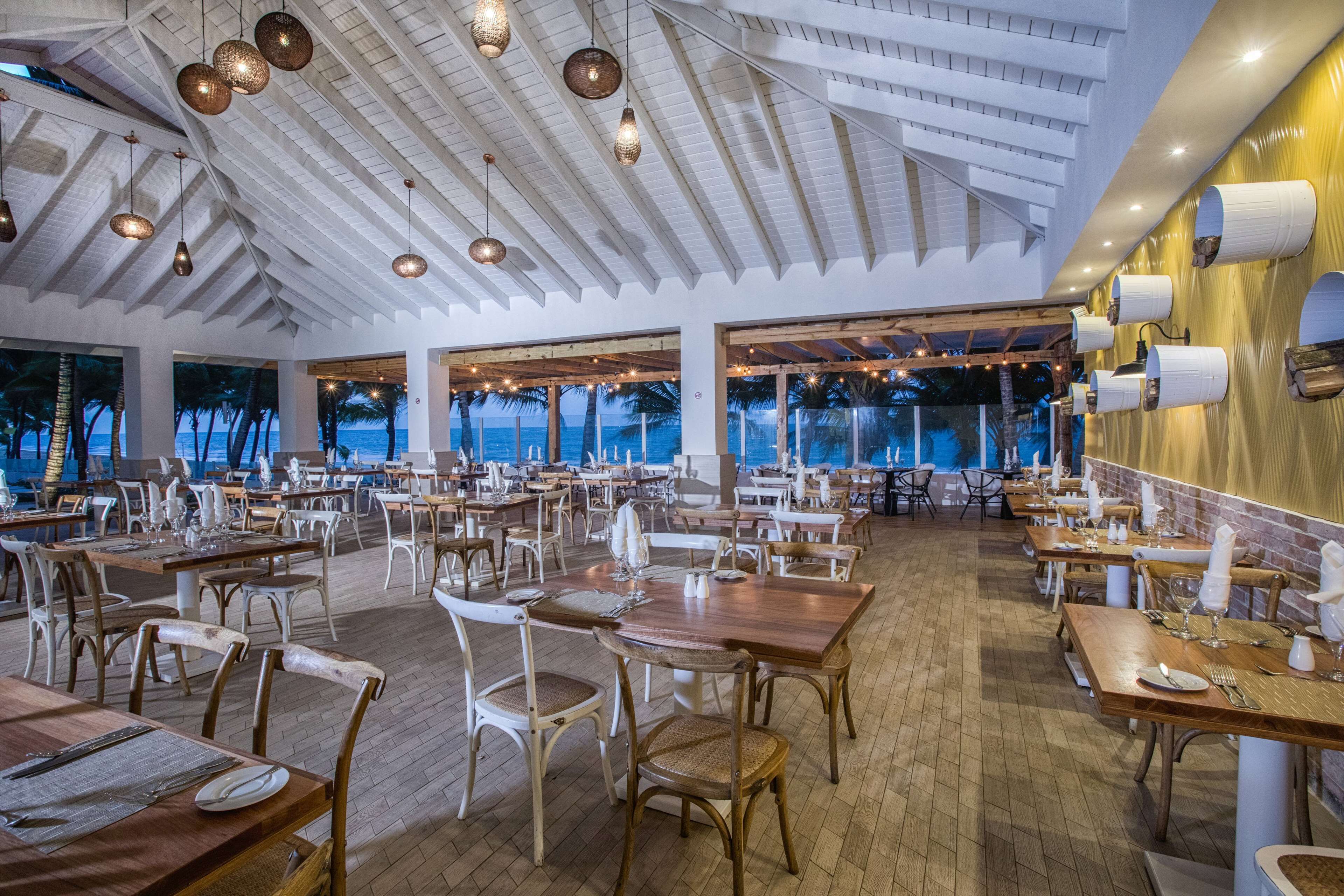 Um restaurante espaçoso com pé-direito alto, mesas e cadeiras de madeira, com vista para o mar.