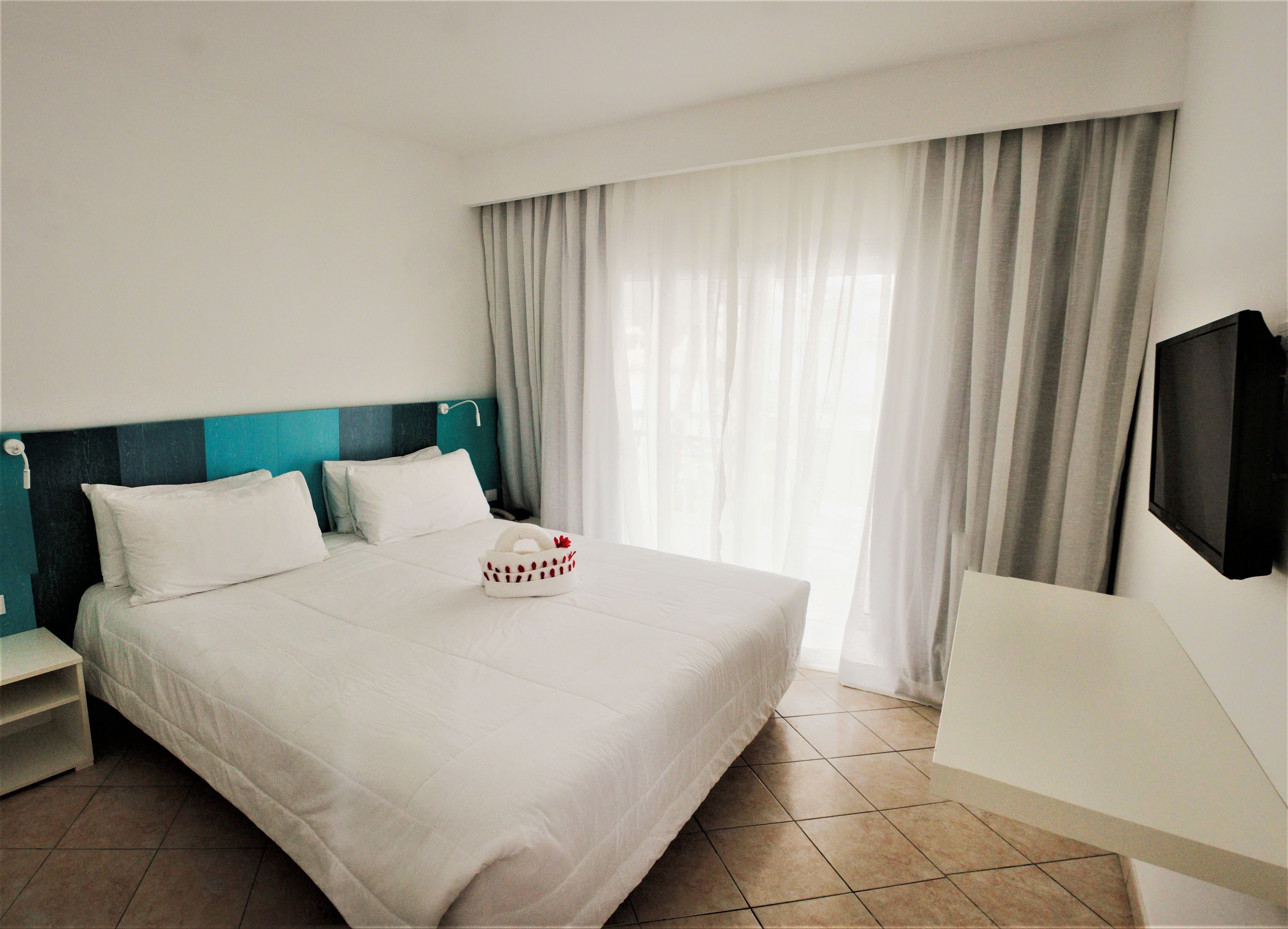 Habitación de hotel con una cama grande, un televisor montado en la pared y una ventana con cortinas.