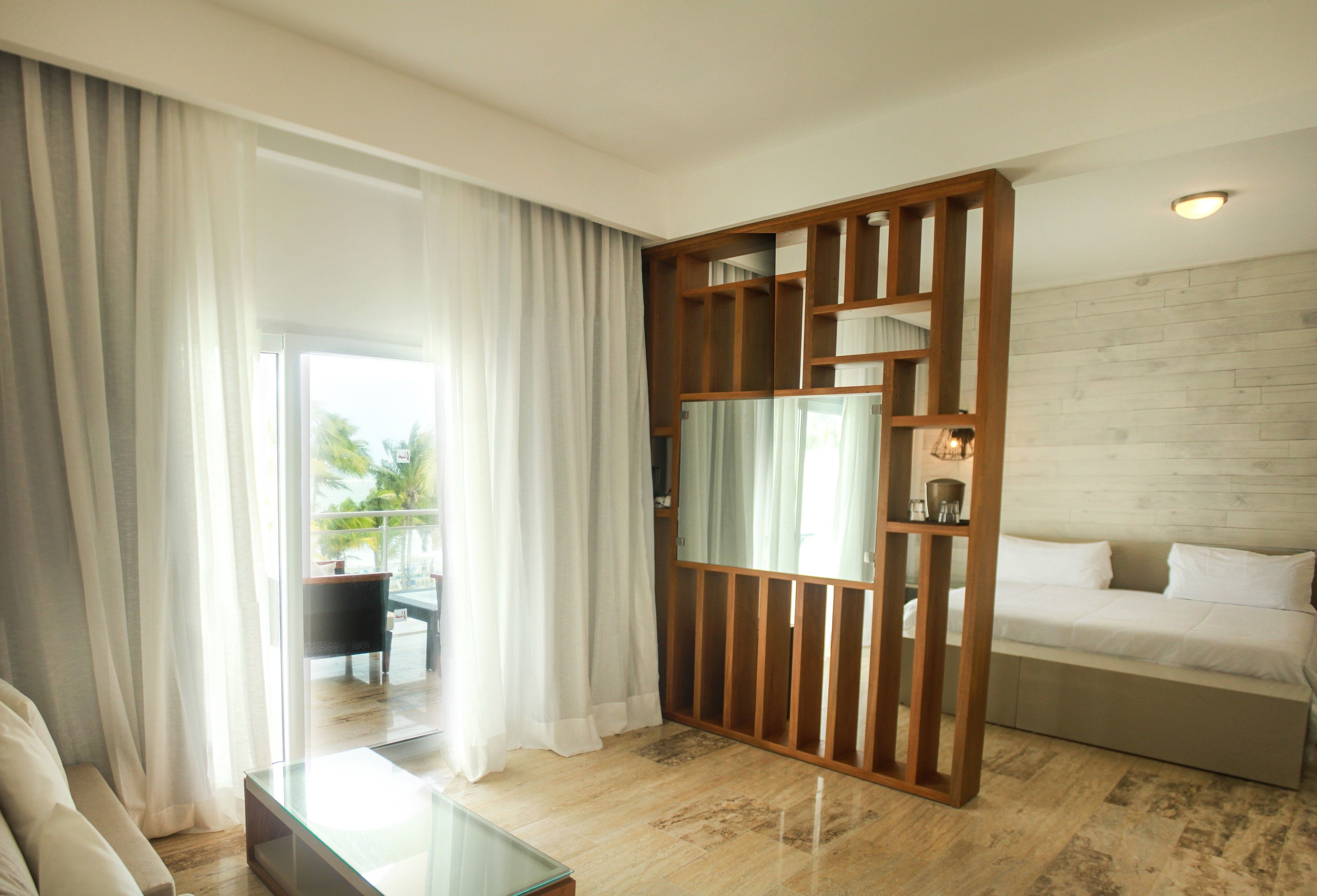 A hotel room with a bed, a glass coffee table, a wooden partition, and a view of the outdoors.