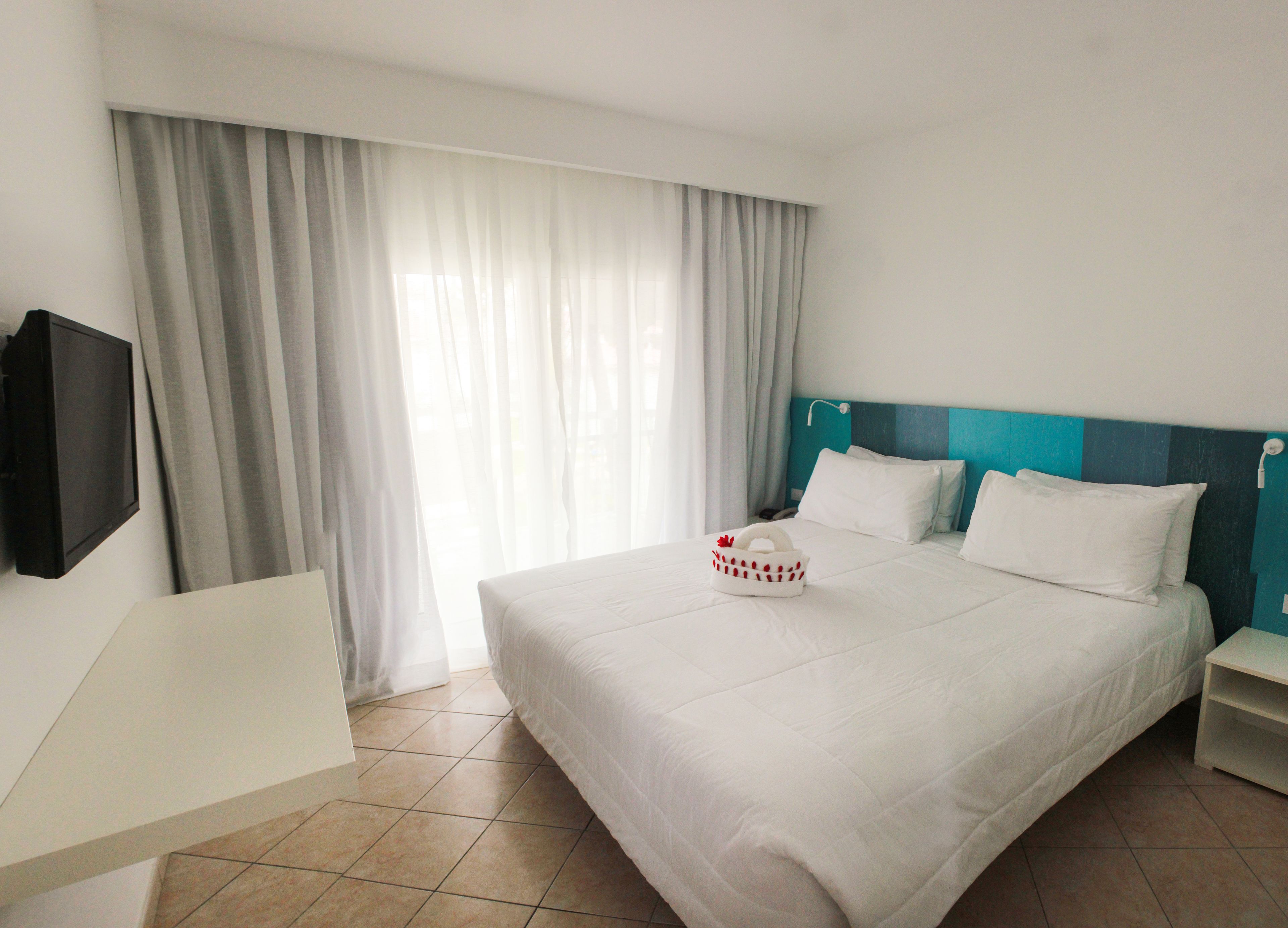 Een hotelkamer met een groot bed, een flatscreen televisie aan de muur en een raam met dunne gordijnen.
