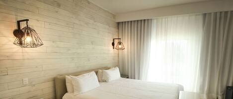 A modern hotel room with a wooden wall, a large bed with white bedding, two wall-mounted light fixtures, a bedside table with a phone and remote control, and a large window with curtains.