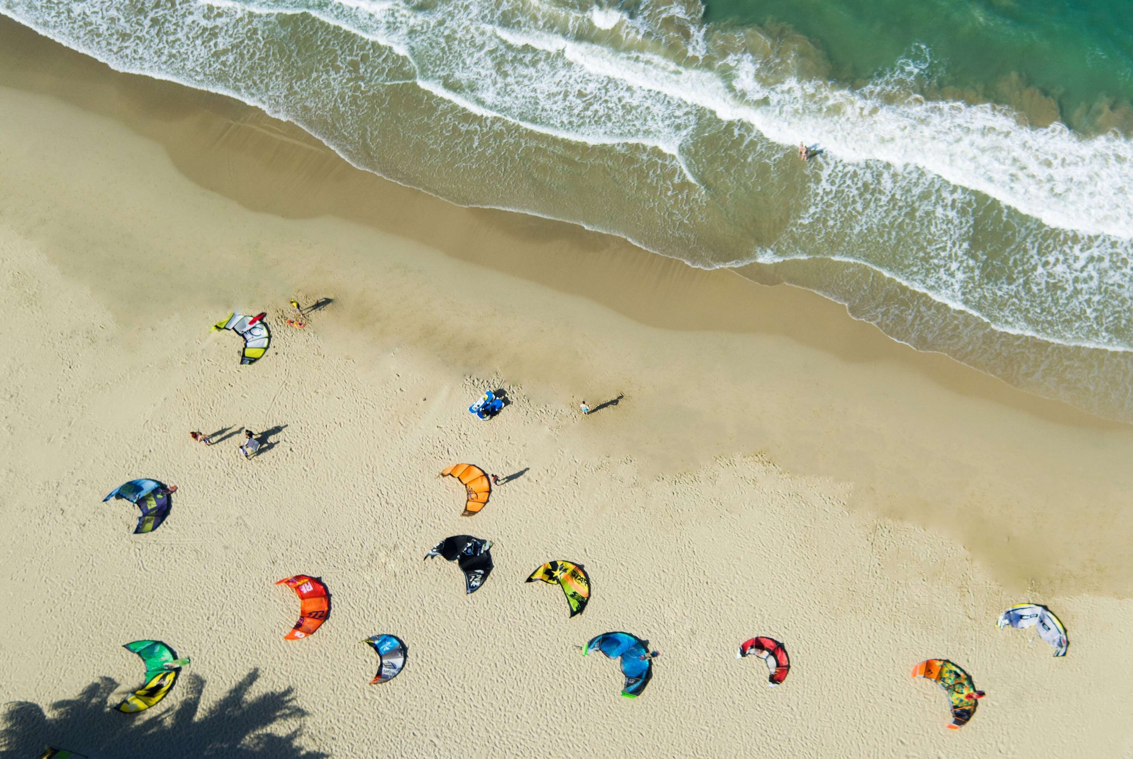 An aerial view of a beach with multiple colorful parasails and people.