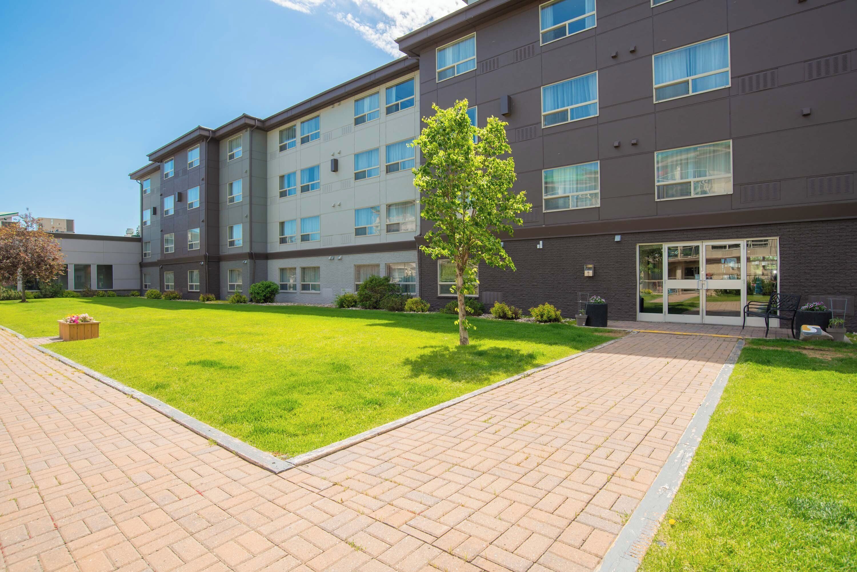 A multi-story hotel with a paved courtyard, a small tree, and a well-maintained lawn.