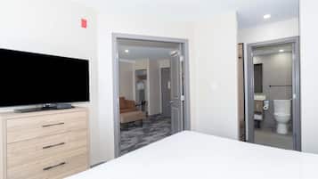A hotel room with a bed, a TV on a dresser, a door leading to a bathroom, and a hallway with a sofa and a coat rack.