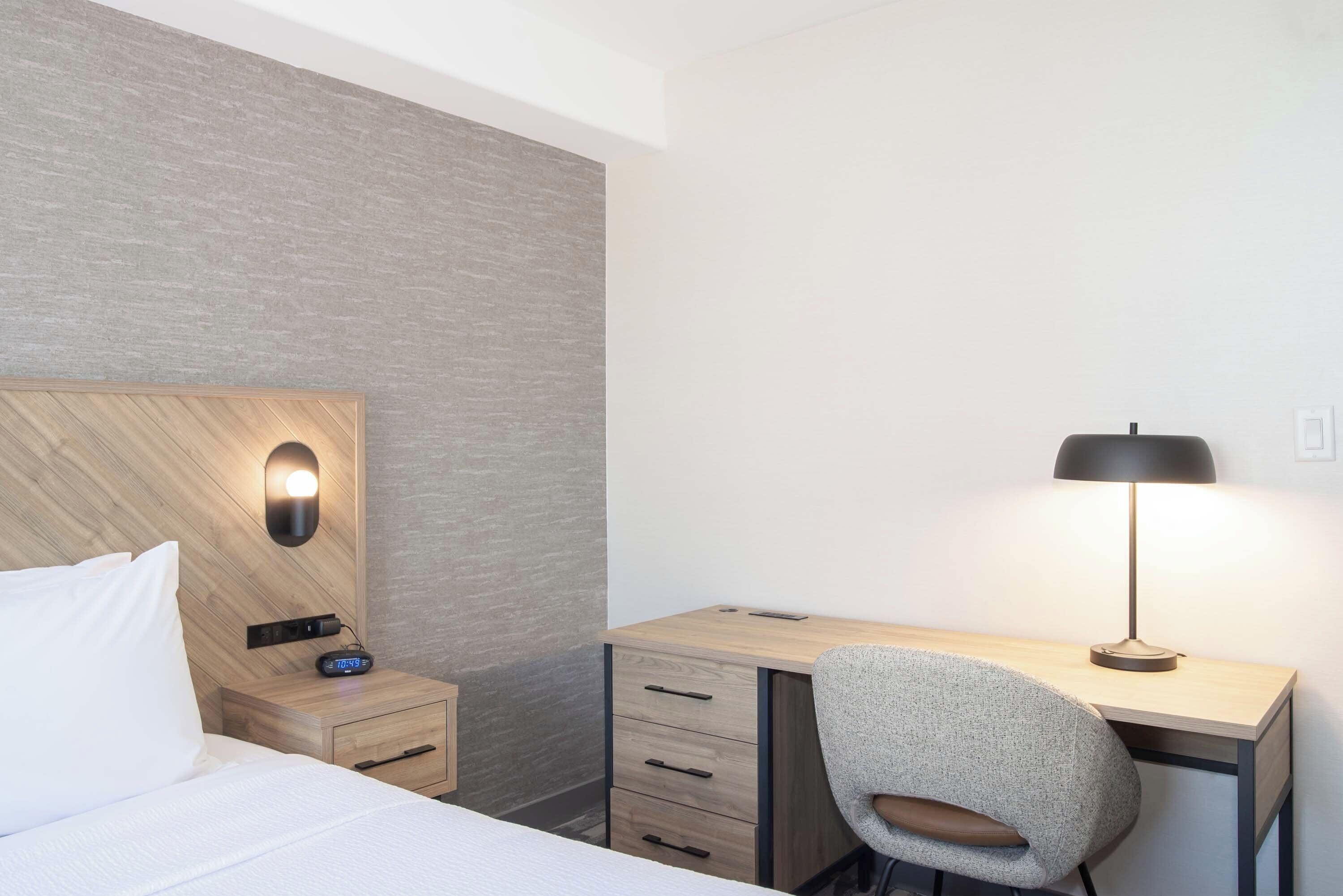 Habitación de hotel con cama, escritorio, silla, lámpara y mesita de noche con radio.