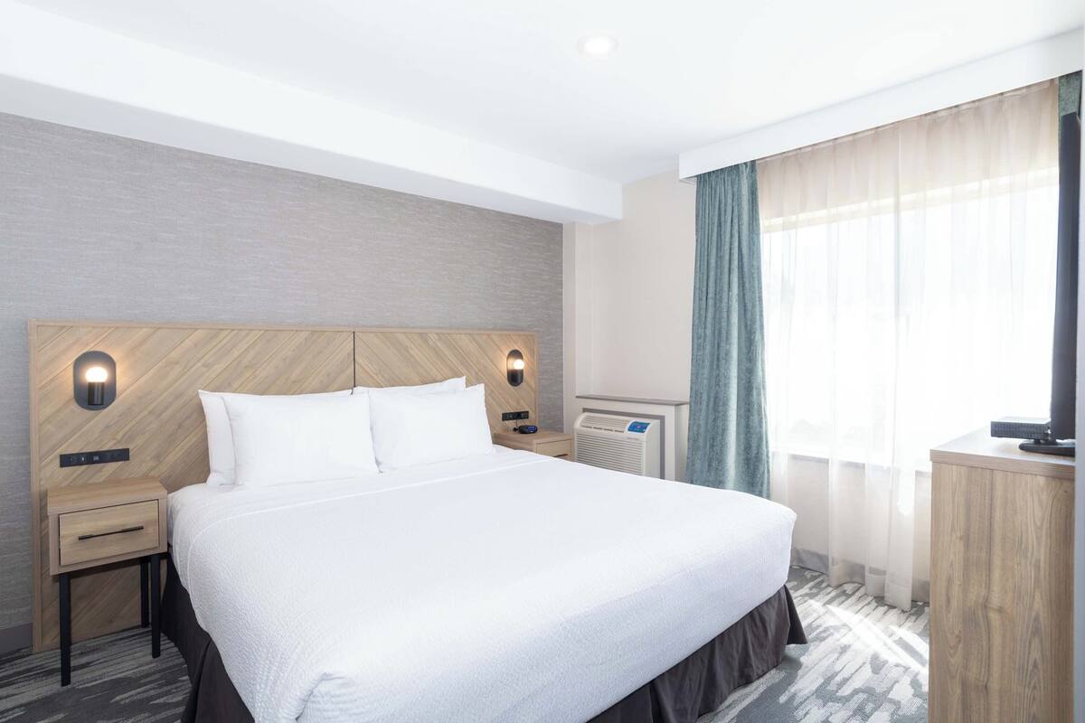 A hotel room with a large bed, two bedside tables, a wall-mounted heater, a television, and a window with sheer curtains.
