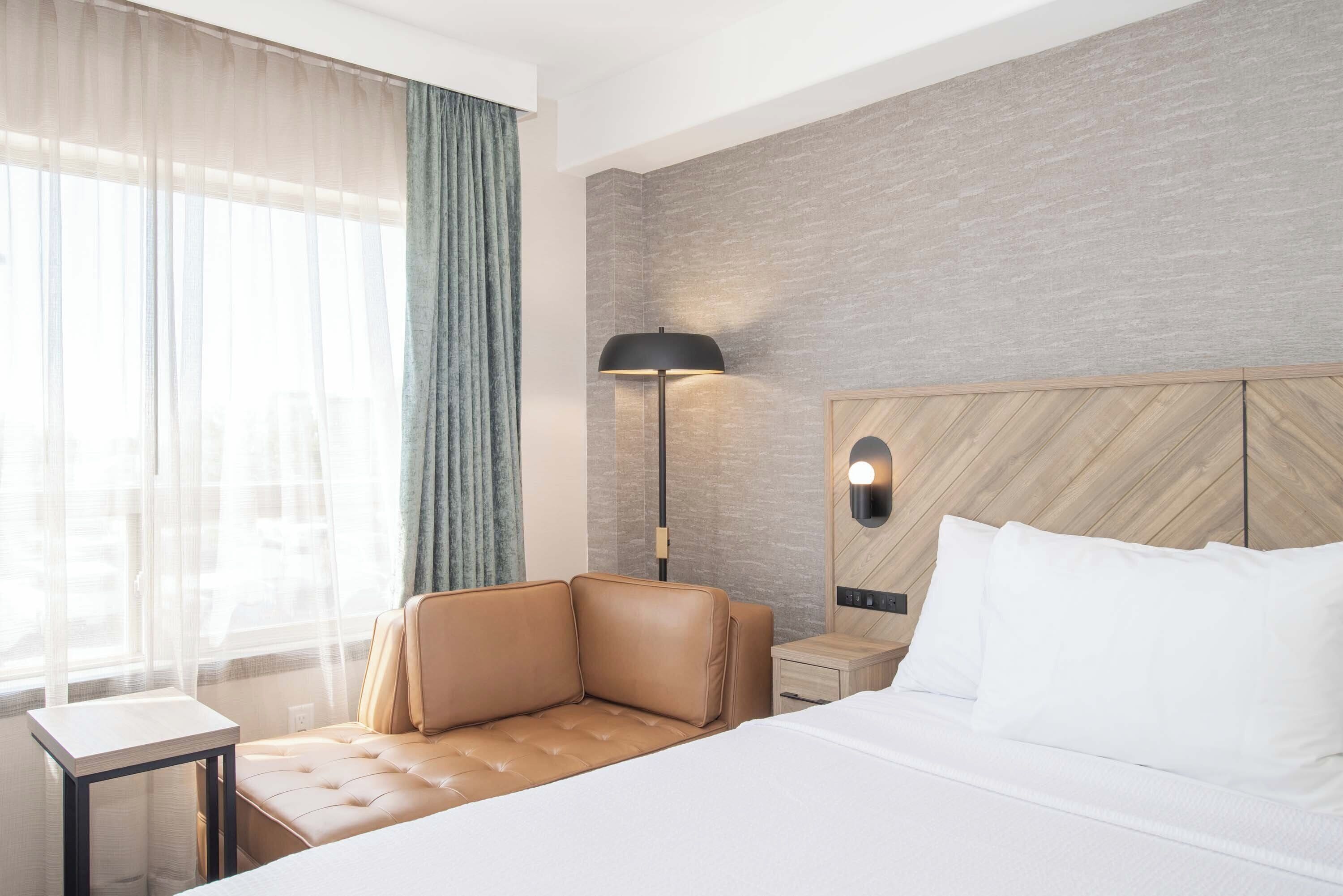 Hotelkamer met een bed, een slaapbank, een nachtkastje, een lamp en uitzicht buiten door dunne gordijnen.