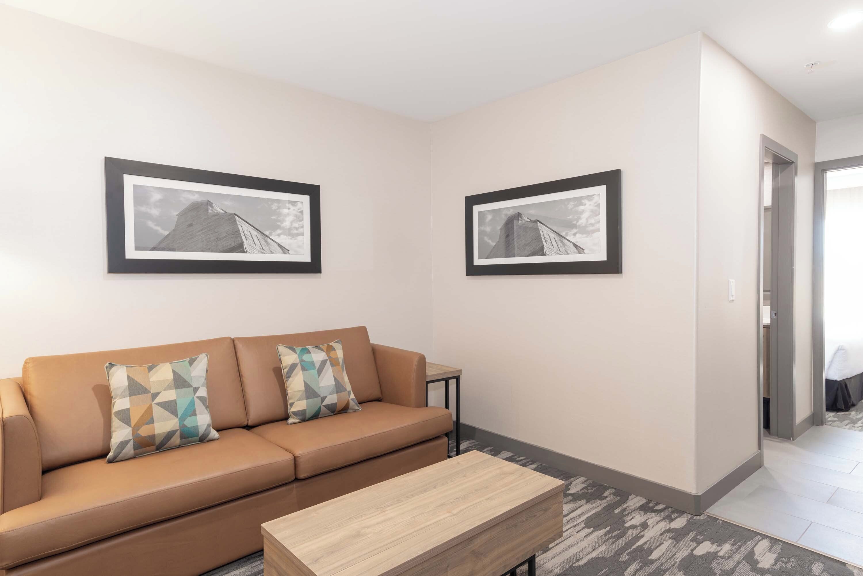 A modern living room with a beige sofa, two patterned cushions, a wooden coffee table, and two framed black-and-white photographs on the walls.