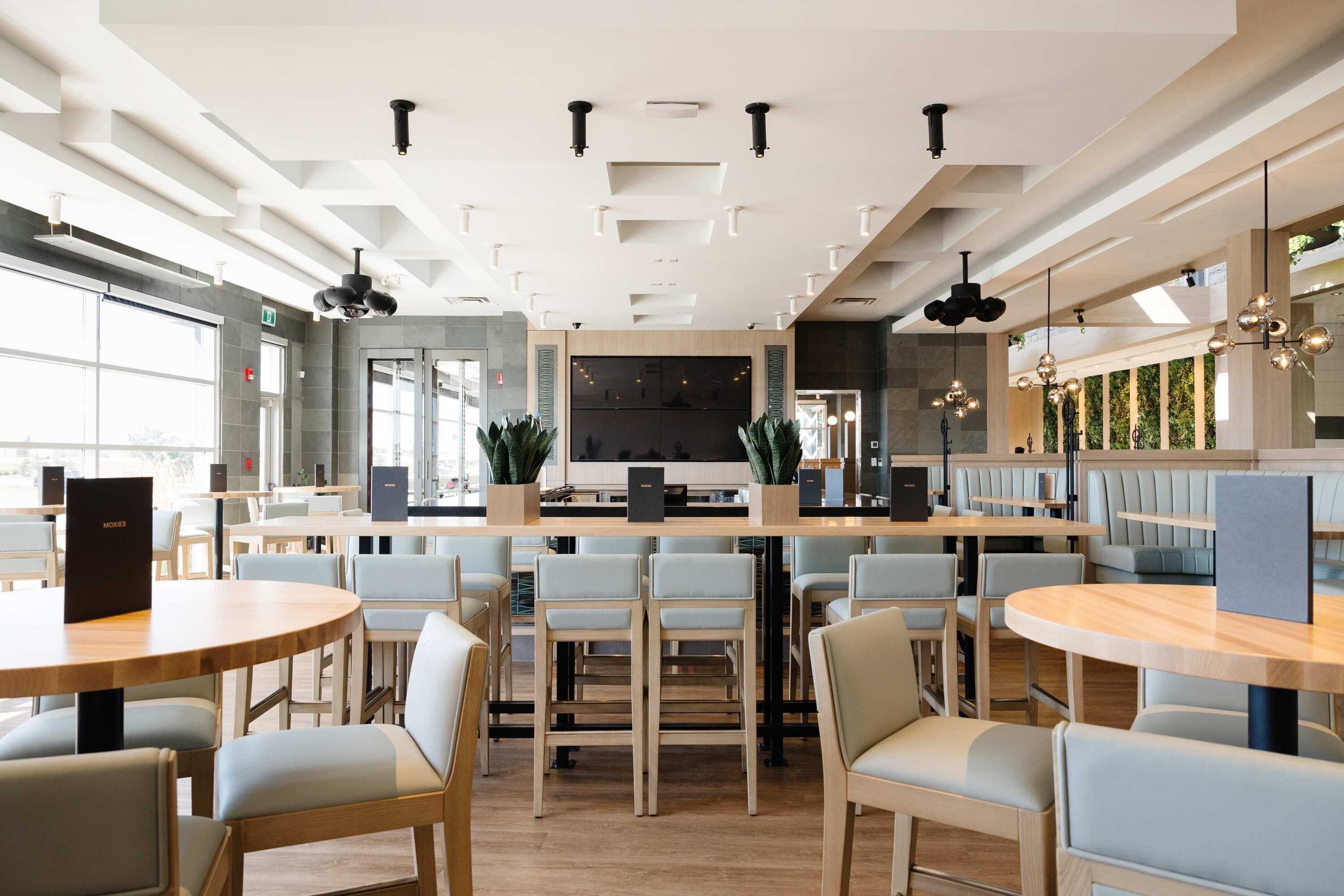 Ein modernes Restaurant mit runden Tischen, hellen Stühlen, einer zentralen Theke und Blick ins Freie.