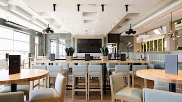 Ein modernes Restaurant mit runden Tischen, hellen Stühlen, einer zentralen Theke und Blick ins Freie.