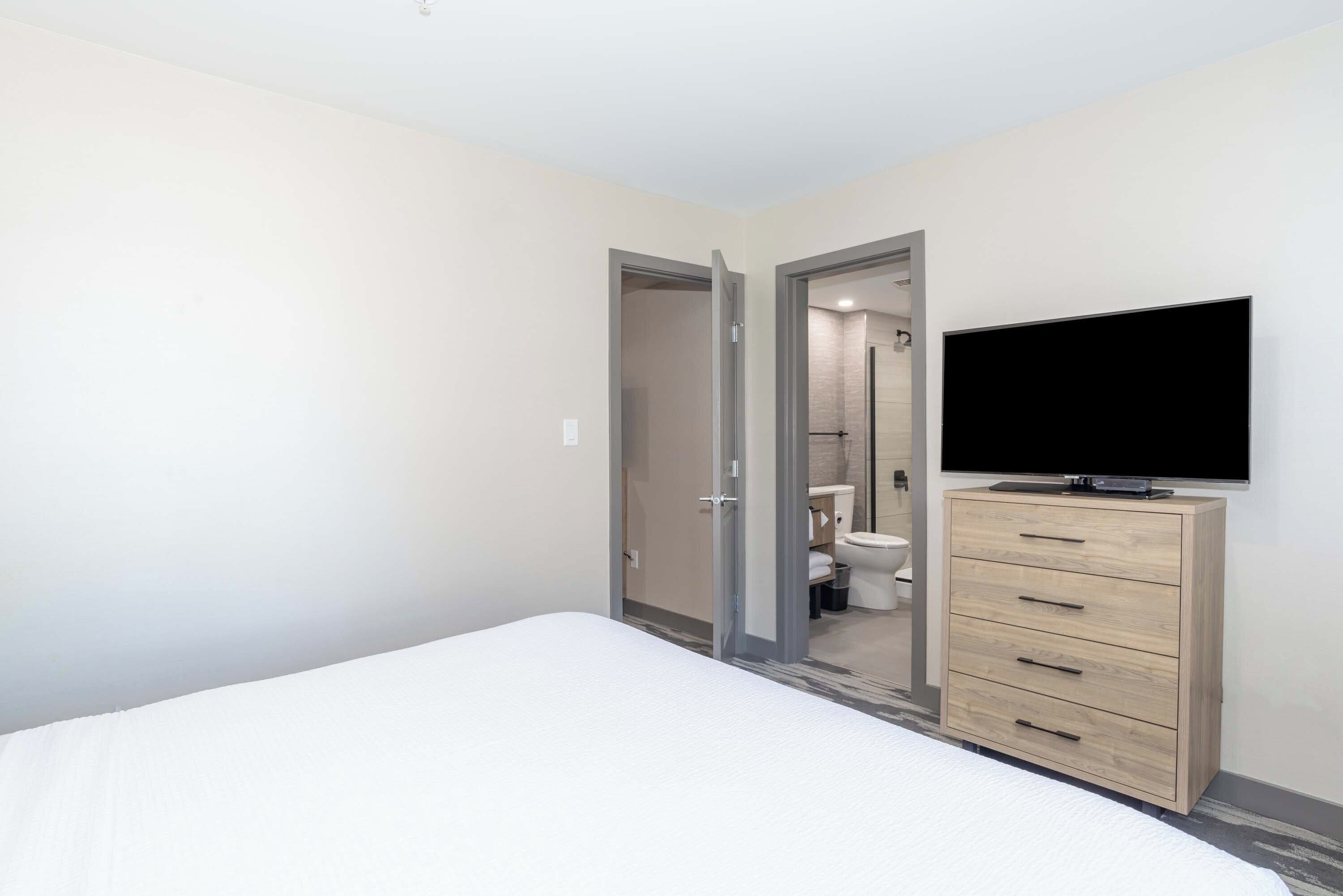 Ein Schlafzimmer mit einem Bett, einem Fernseher auf einer Holzkommode und einem durchgegangenen Tür erkennbaren Badezimmer.
