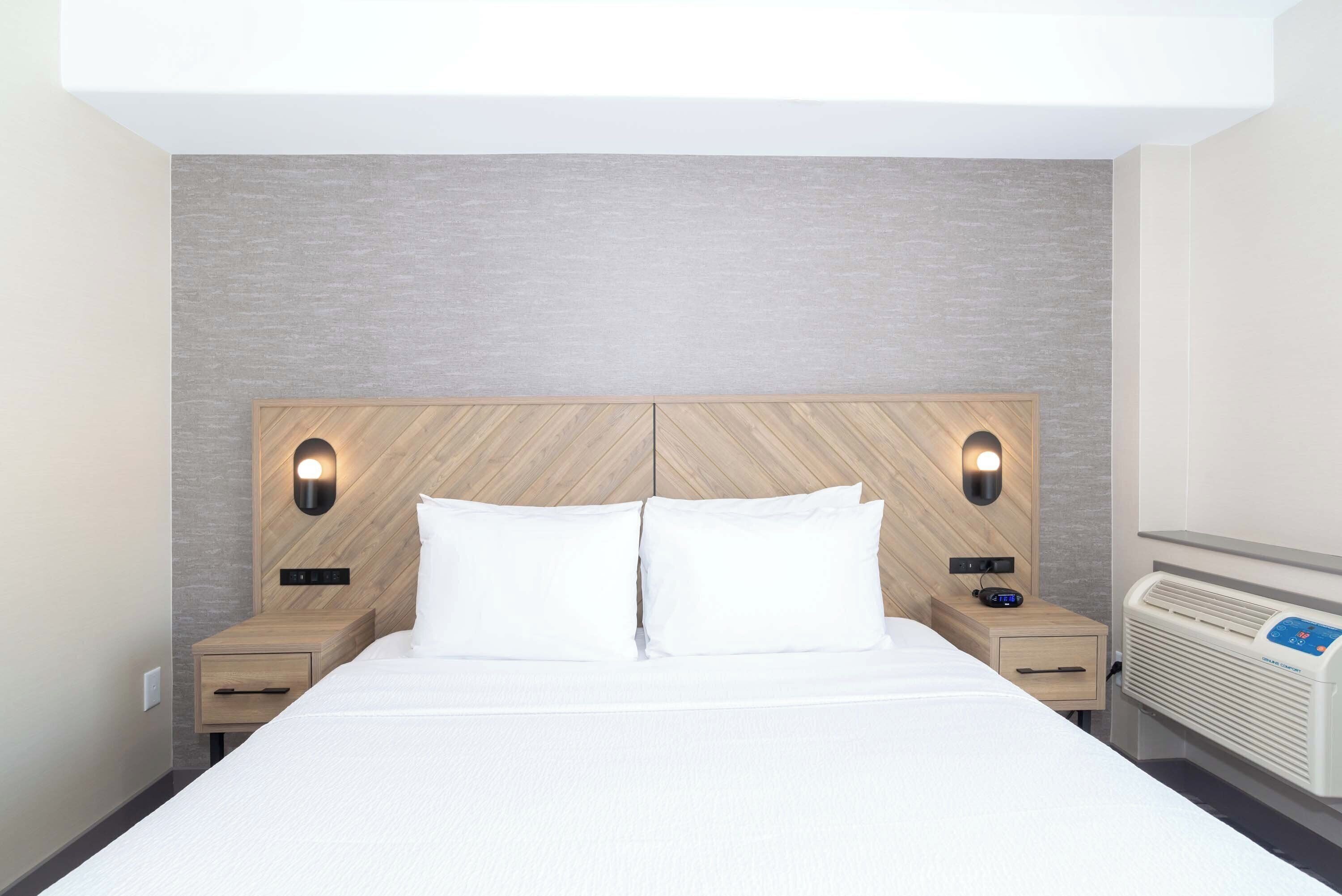 A modern hotel room with a large bed, two bedside tables, a built-in heater, and wall-mounted lighting.