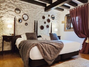 A bedroom with a stone wall, a bed with a brown blanket, two bedside tables with lamps, and a window with curtains. - Hotel Casa del Marqués (Santillana del Mar)