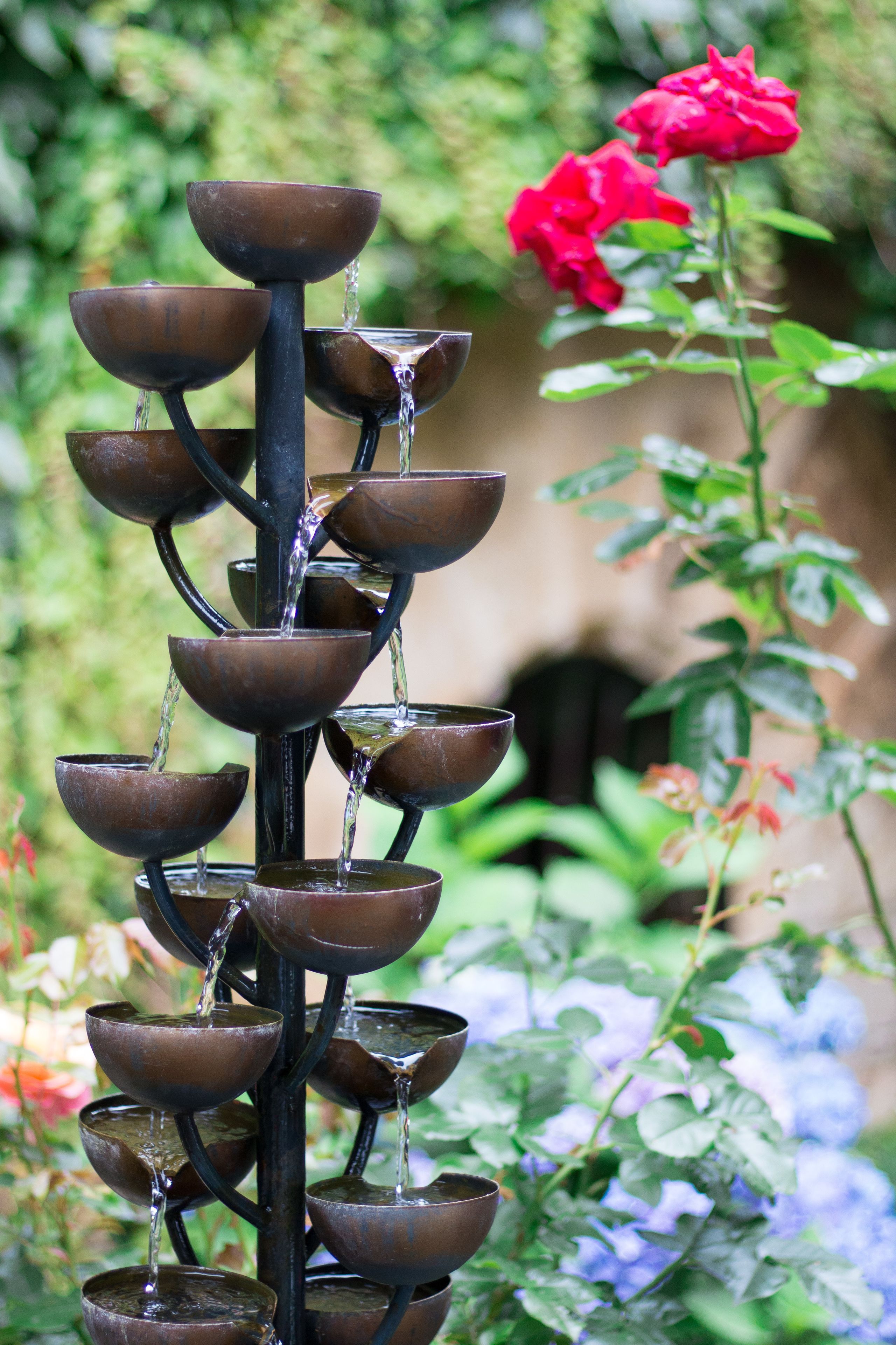 A tiered water feature with multiple bowls, surrounded by lush greenery and a red rose.