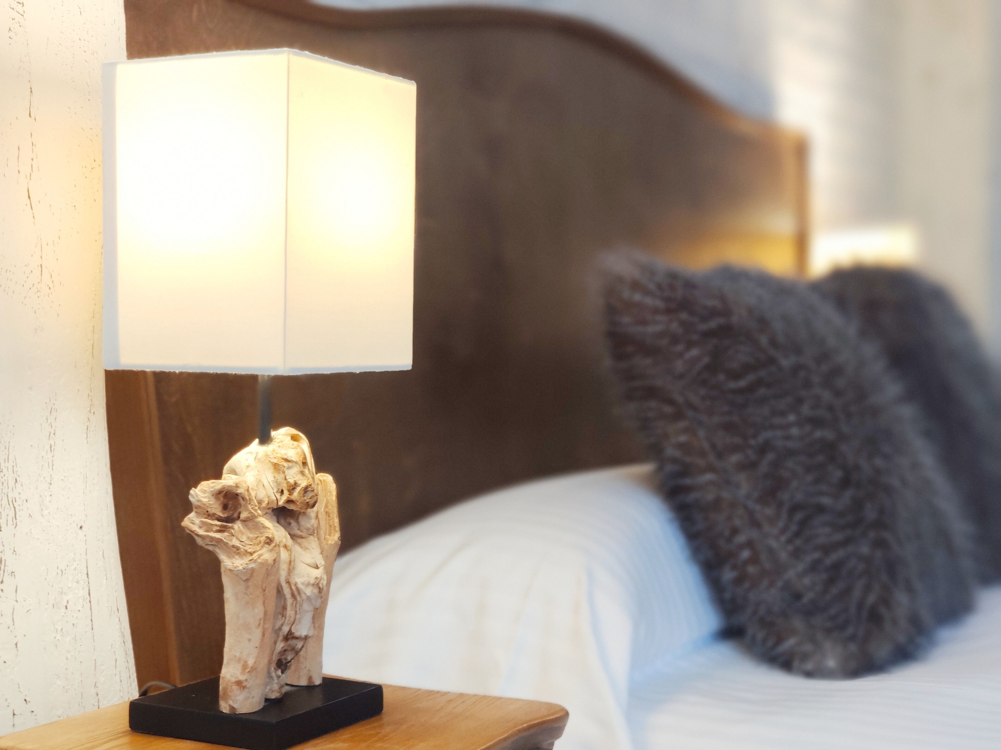 Een nachtkastje met een lamp en een decoratief object, een bed met witte lakens en een donker kussen, en een houten hoofdeinde.