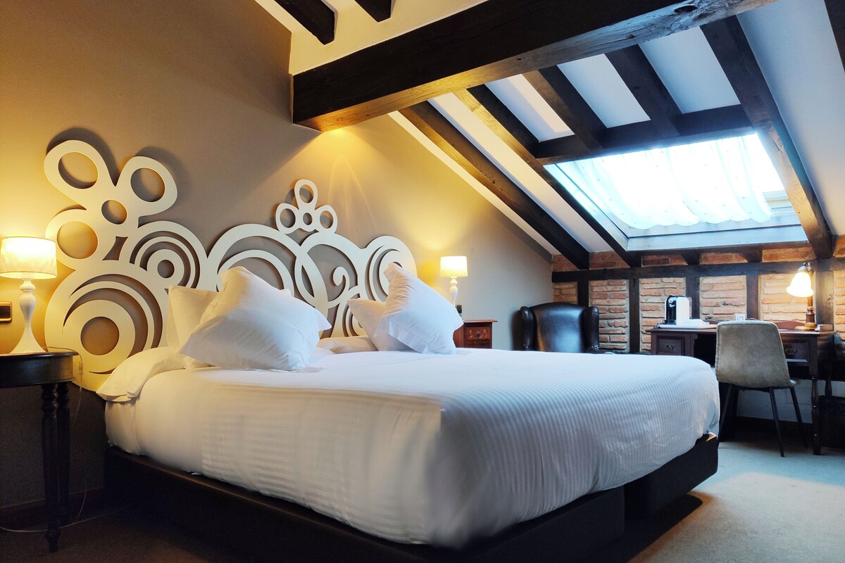 Una cama bien tendida con sábanas y almohadas blancas, un cabecero decorativo, un escritorio con silla y un tragaluz arriba.