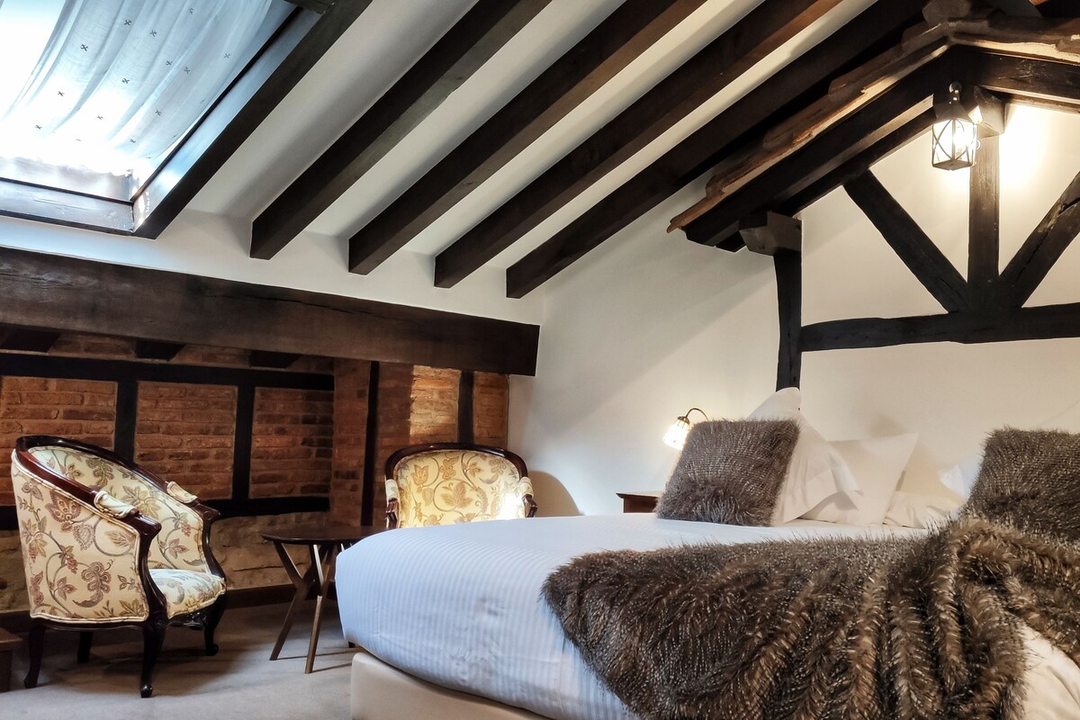 Un dormitorio con una cama, dos sillas, una pared de ladrillo y un techo inclinado con vigas a la vista.