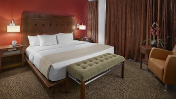 Una habitación de hotel con una cama grande, un cabecero de madera, una mesita de noche con una lámpara, una pequeña mesa con una planta y un sillón de cuero marrón.