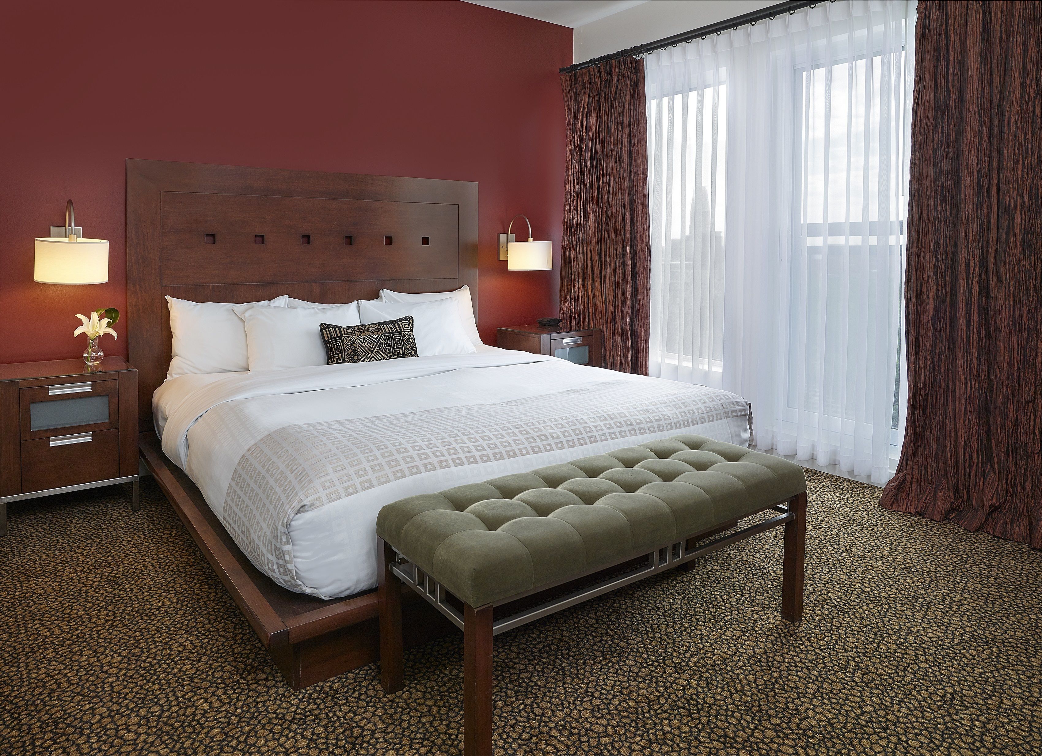 A hotel room with a large bed, two bedside tables, a nightstand, a bench, and a window with curtains.
