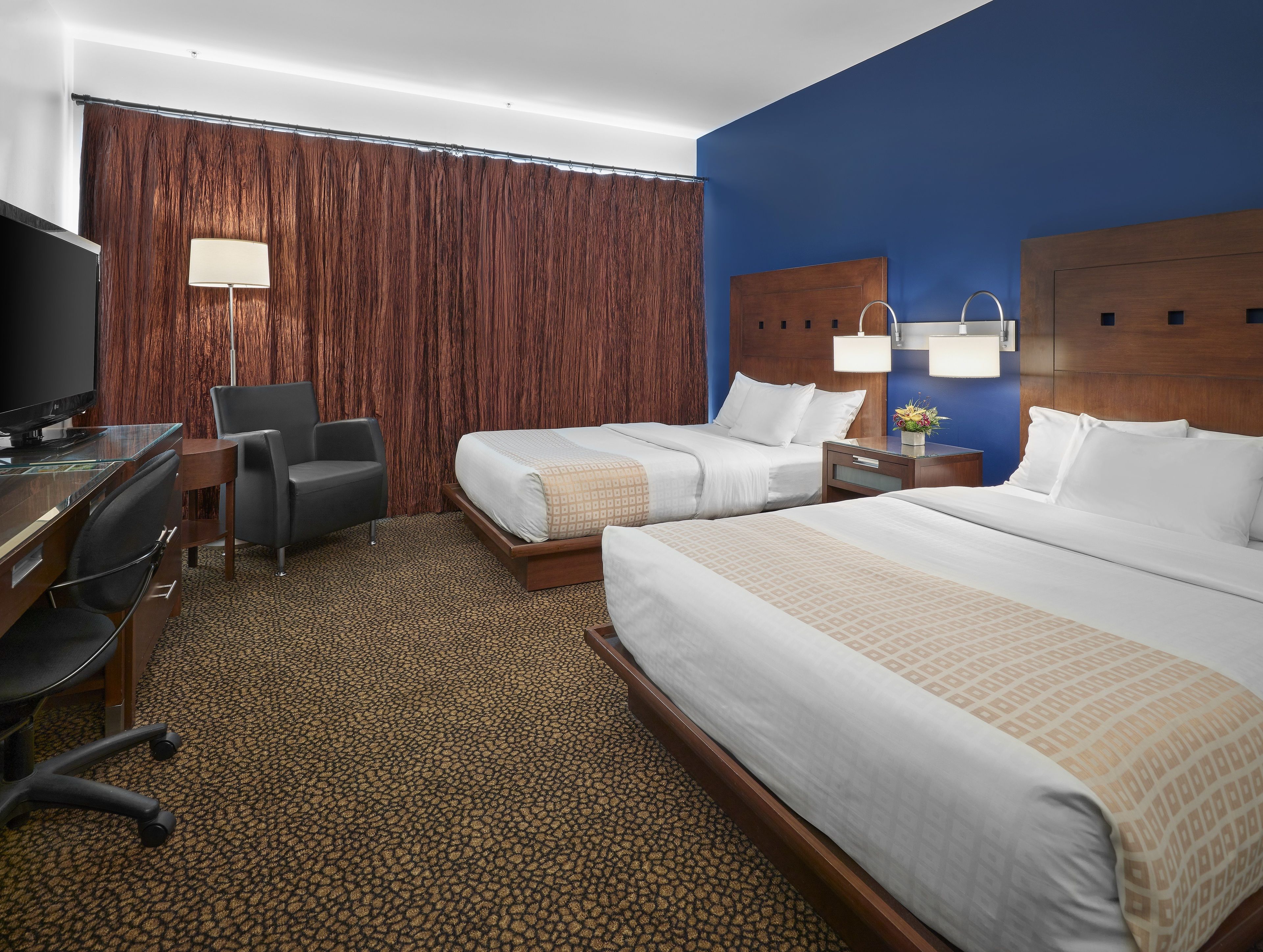 Quarto de hotel com duas camas, uma escrivaninha com televisão, uma cadeira e uma janela grande com cortinas.