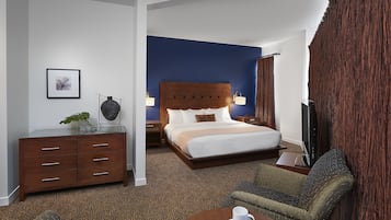 Habitación de hotel con una cama grande, un escritorio con cajones, una silla, una mesita de centro y una televisión.