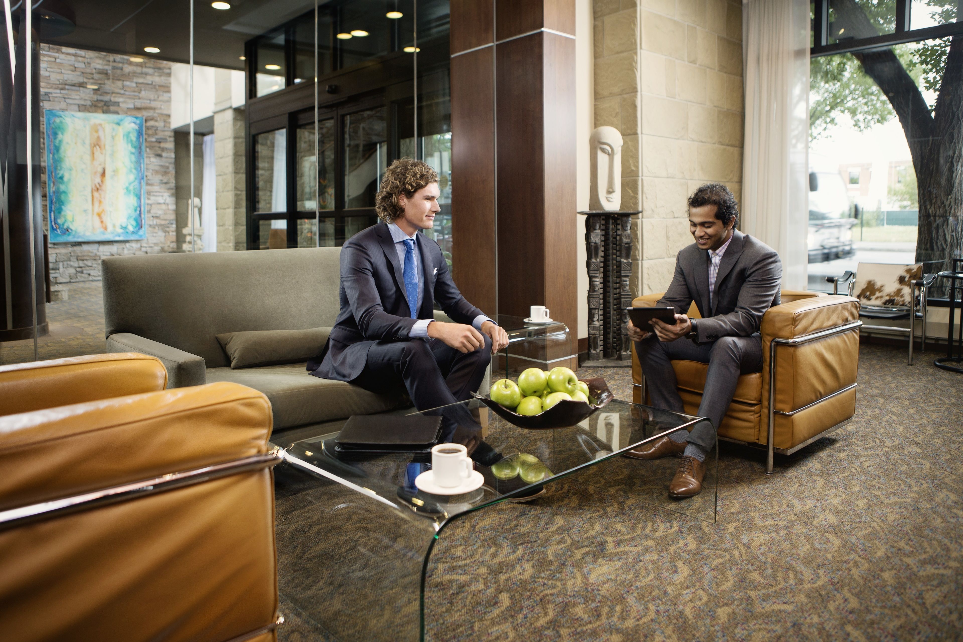 Twee mannen in pak zitten op leren banken in een moderne lobby met een glazen salontafel, een laptop en een schaal appels.