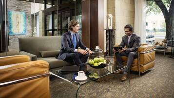 Twee mannen in pak zitten op leren banken in een moderne lobby met een glazen salontafel, een laptop en een schaal appels.