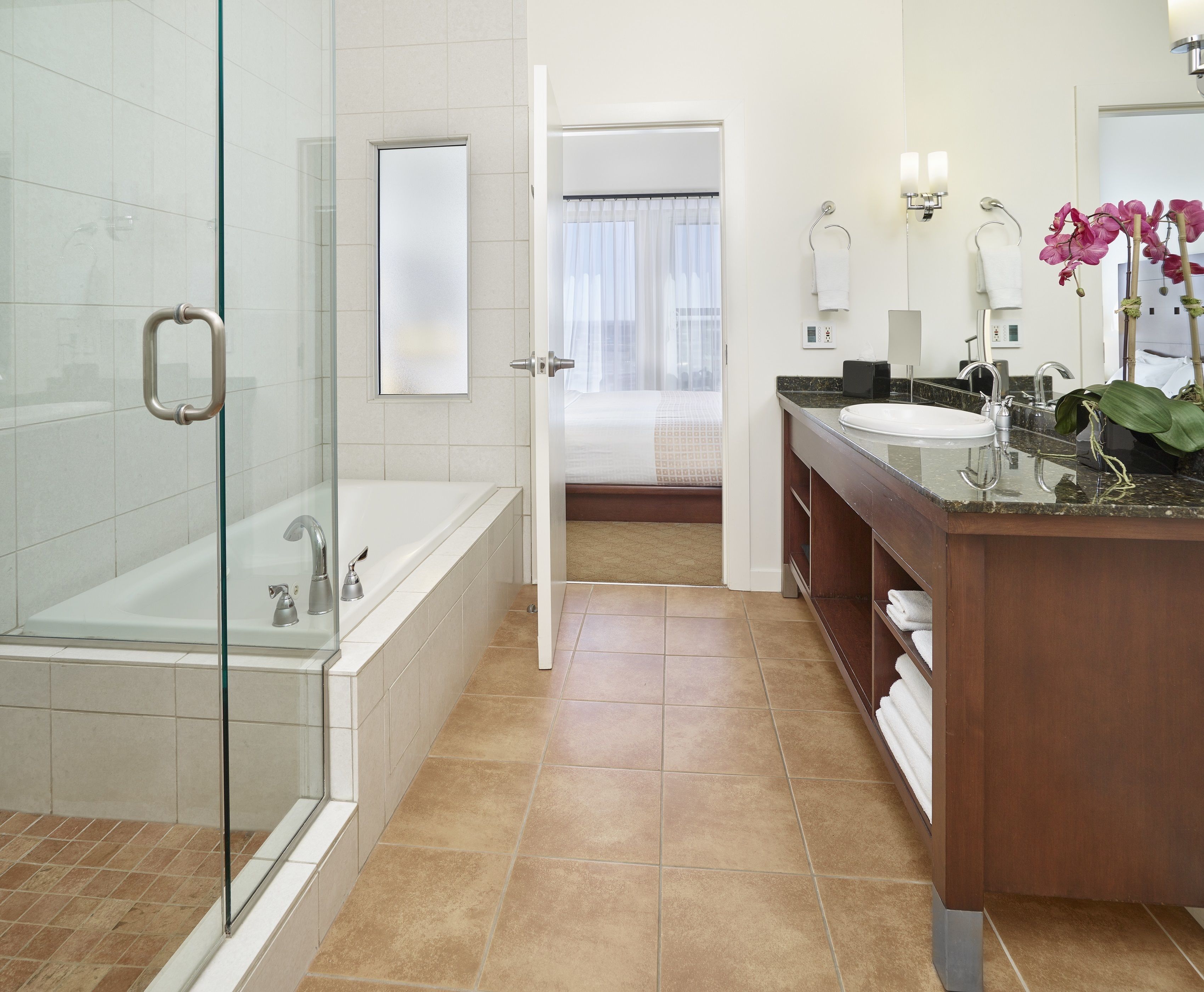 Un baño moderno con ducha de vidrio, una bañera independiente, un tocador con lavabo y espejo, y una habitación visible con cama y cortinas.