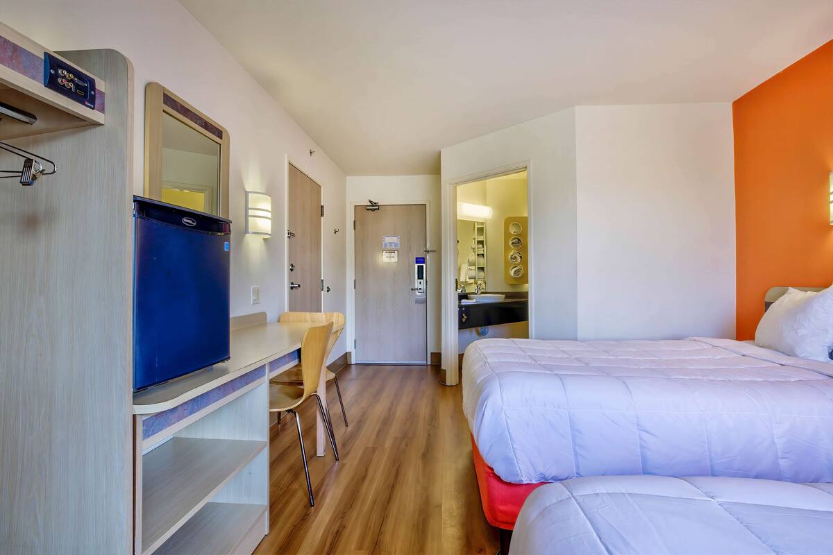 Une chambre d’hôtel avec un lit, un bureau, une chaise, une armoire et une salle de bain visible à travers une porte ouverte.