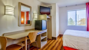 Une chambre d’hôtel avec un bureau en bois, deux chaises, une télévision, un lit recouvert d’une couette blanche et une fenêtre avec des rideaux.