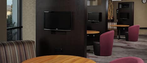 A modern hotel lobby with round wooden tables, comfortable seating, and a large TV screen.