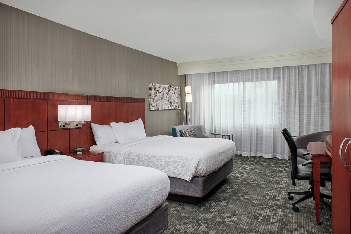 A hotel room with two beds, a desk, a chair, a lamp, and a window with curtains.
