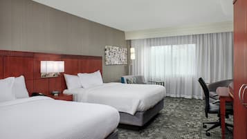 Habitación de hotel con dos camas, un escritorio, una silla, una lámpara y un ventanal con cortinas.