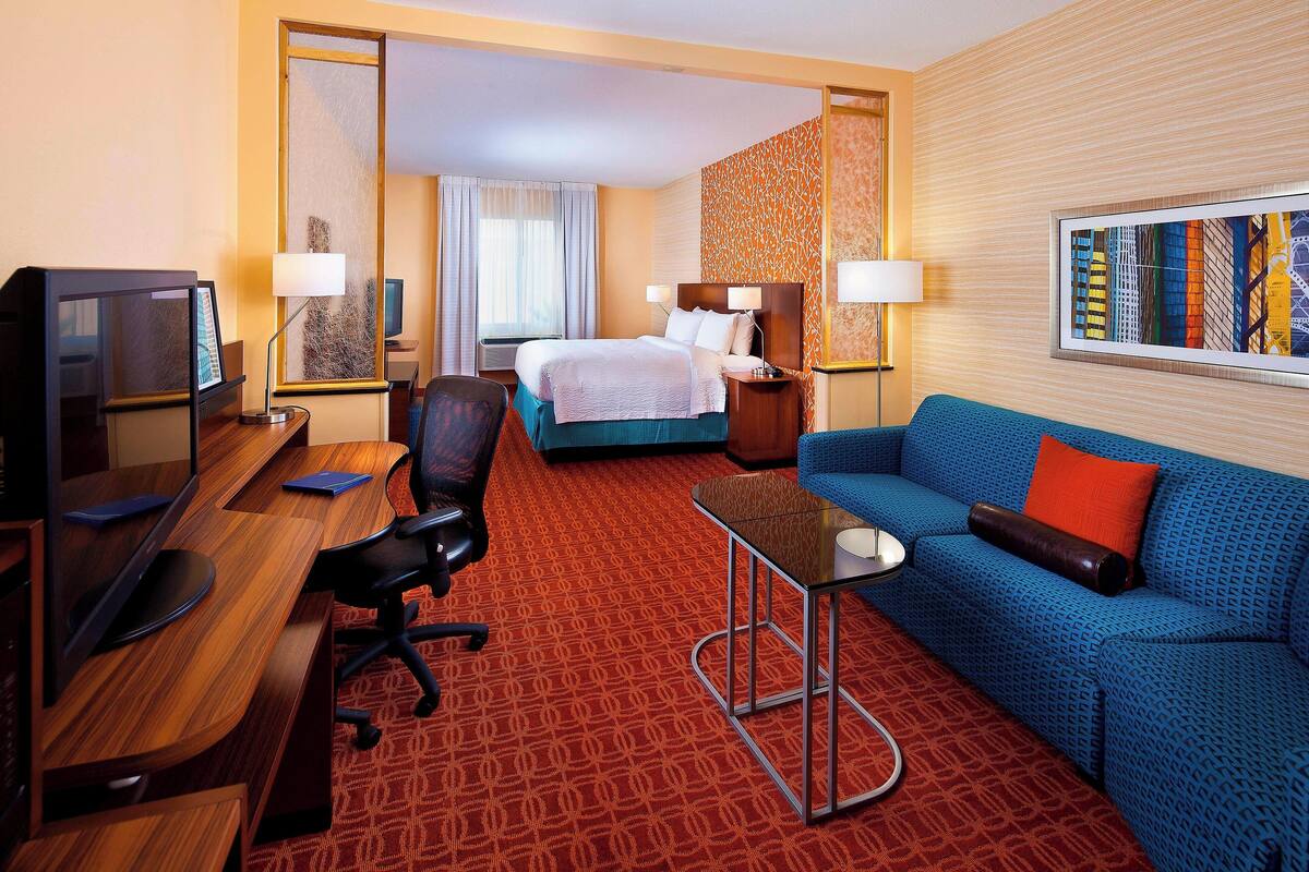 A hotel room with a bed, a desk with a computer, a blue sofa, and a large window.