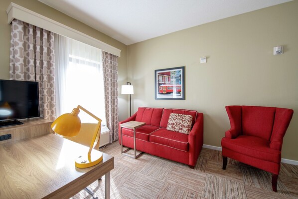 A living room with a red sofa, a red armchair, a wooden desk with a yellow lamp, a television, and a patterned rug. - Hampton Inn & Suites Lafayette (Lafayette)