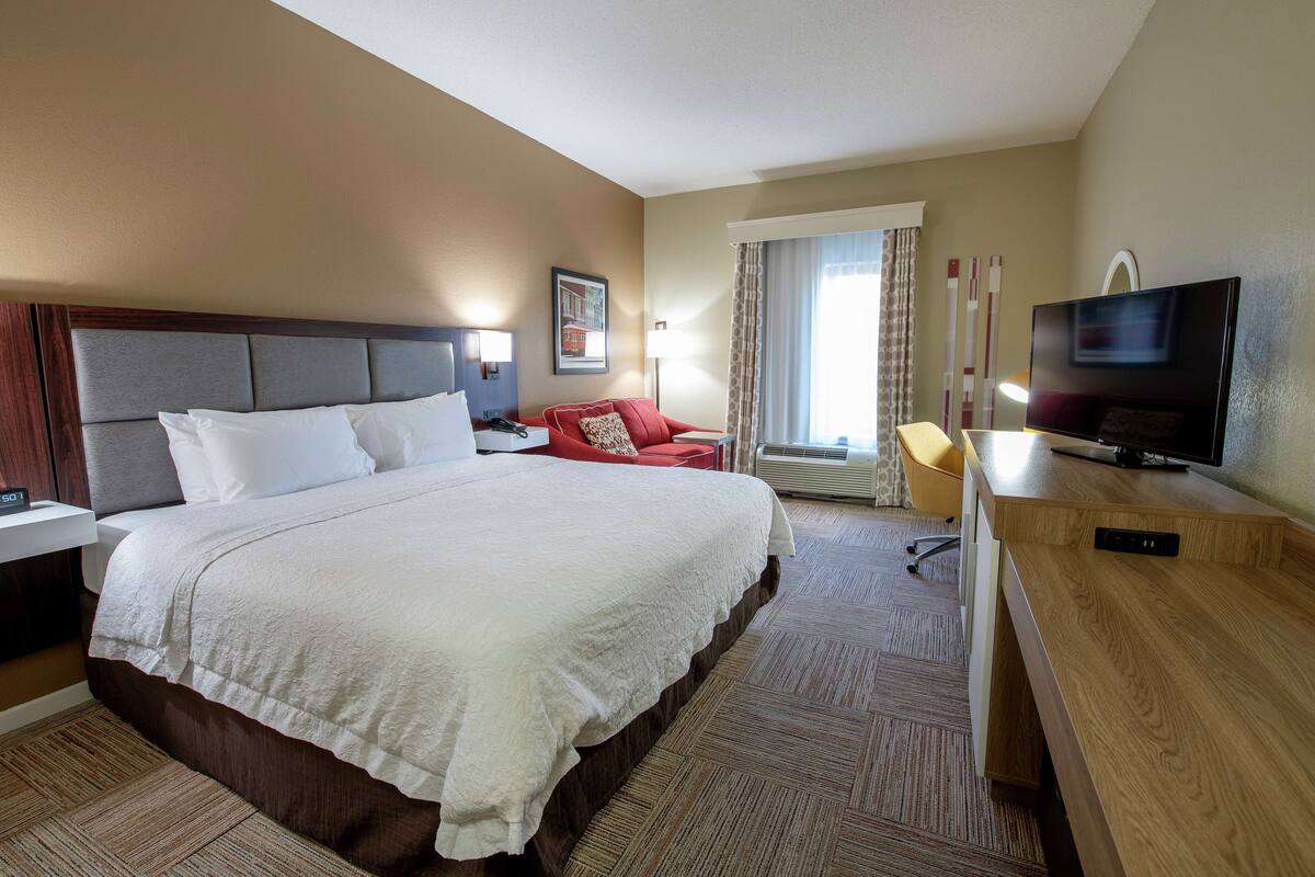 A hotel room with a large bed, a desk with a TV, a chair, and a window with curtains.