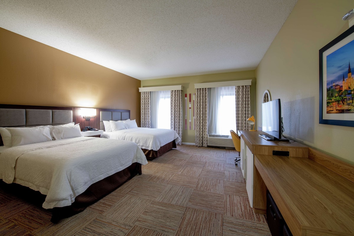 A hotel room with two beds, a desk with a TV, and a large window with curtains.