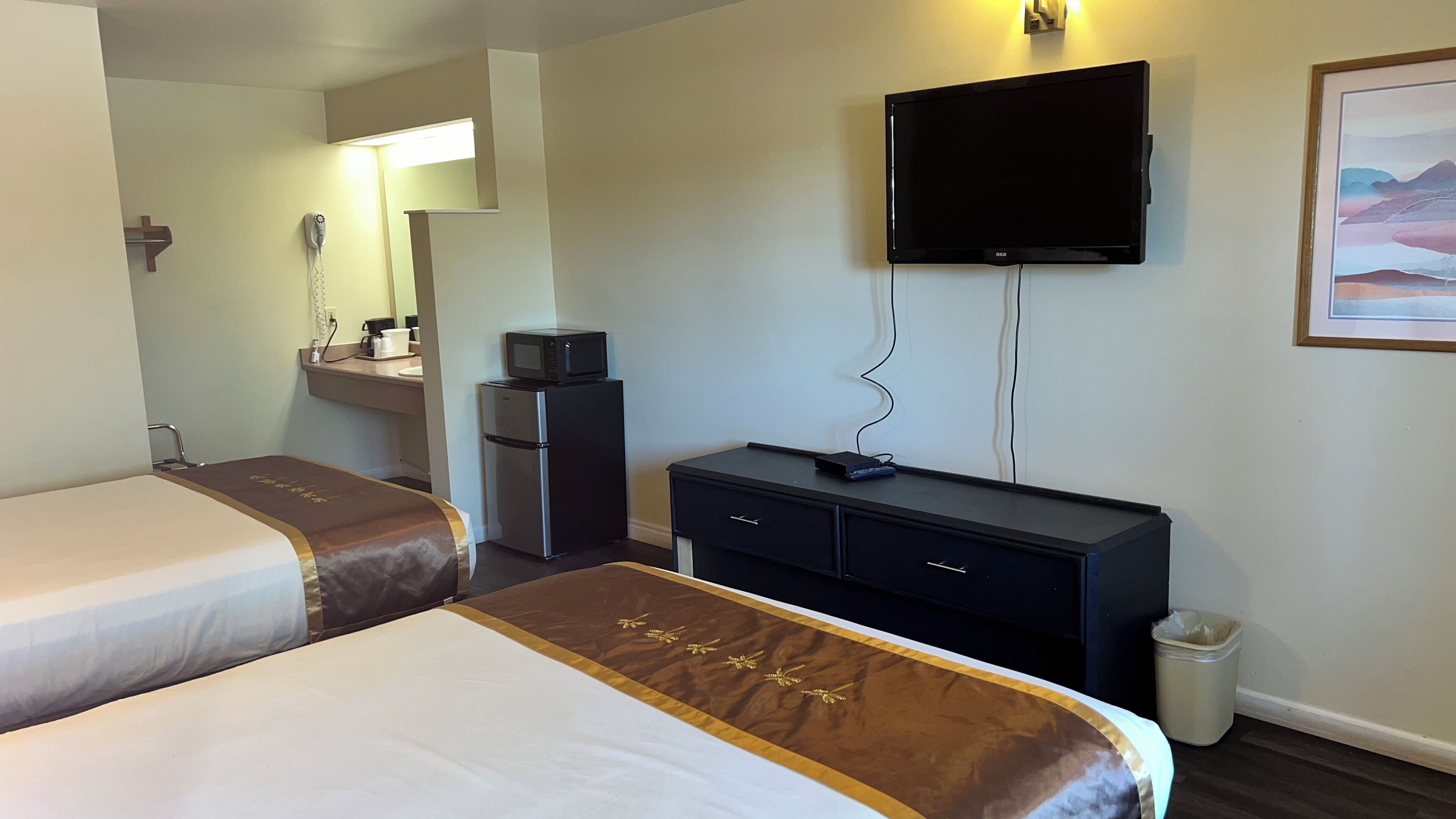 A hotel room with two beds, a small kitchenette, a television, and a painting on the wall.