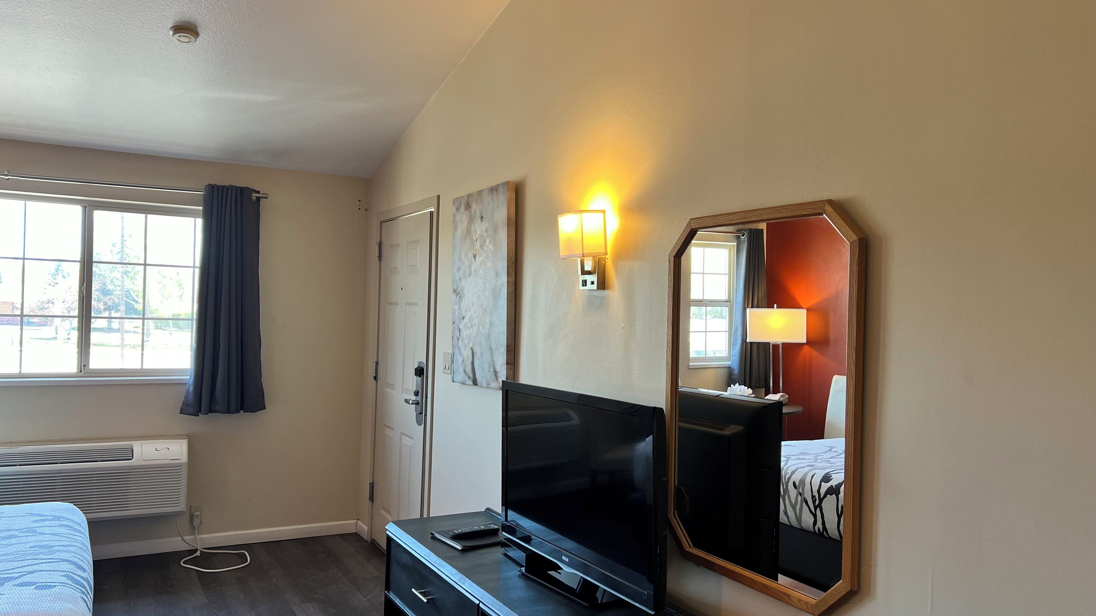 A hotel room with a large flat-screen TV, a bed with a patterned bedspread, a wall-mounted mirror, a door with a light switch, and a window with curtains.