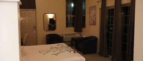 A hotel room with a bed, two chairs, a small table, an air conditioning unit, a window with curtains, and a door.